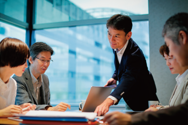 A group of business professionals seated around a conference table. One person stands and gestures towards a laptop, while others lean in to listen and engage. The table is equipped with documents and stationery, and large windows in the background provide a view of an outdoor area.