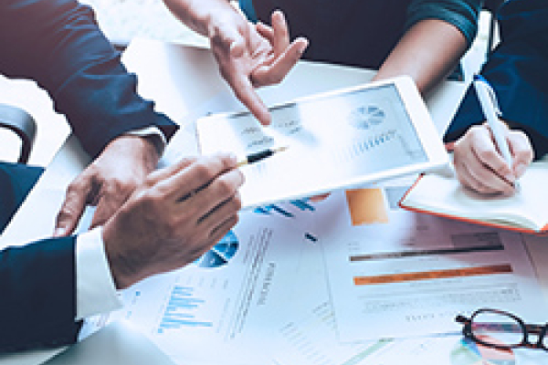 An image capturing a business meeting atmosphere where individuals are discussing data and strategies. Hands are shown pointing at a tablet displaying various graphs and charts, while documents with data visuals and notes are spread across the table. One person holds a pen, indicating active engagement in the discussion. The setting appears collaborative, highlighting teamwork and analysis.