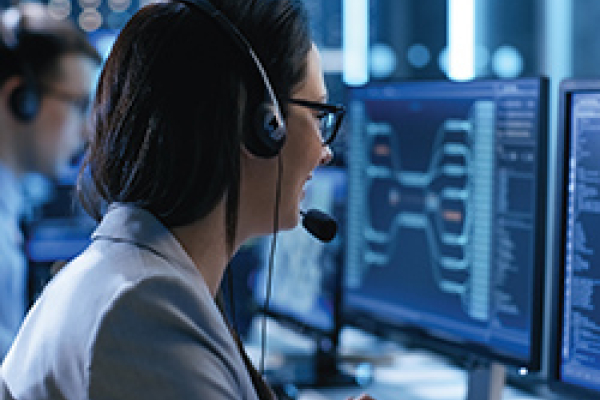 A blurred image showing a person in a professional setting, seated in front of multiple computer monitors. The individual is wearing a headset and appears to be engaged with the screens, which display various data visualisations and code. The background suggests a high-tech environment, possibly in a control room or tech support centre.
