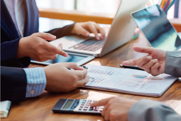 Hands are engaged in discussion over a document featuring charts and graphs on a wooden table. Laptops and a calculator are visible, indicating a collaborative workspace. One hand points to the document, while another gestures in conversation.