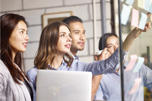 一群專業人員在工作場所協作,積極參與討論。一個人指著貼滿便利貼的玻璃板,而另一個人似乎正在做筆記或使用筆記型電腦。氣氛顯示此為動態腦力激盪課程,從以使用者為中心的角度專注於進階設計和視覺化技術。